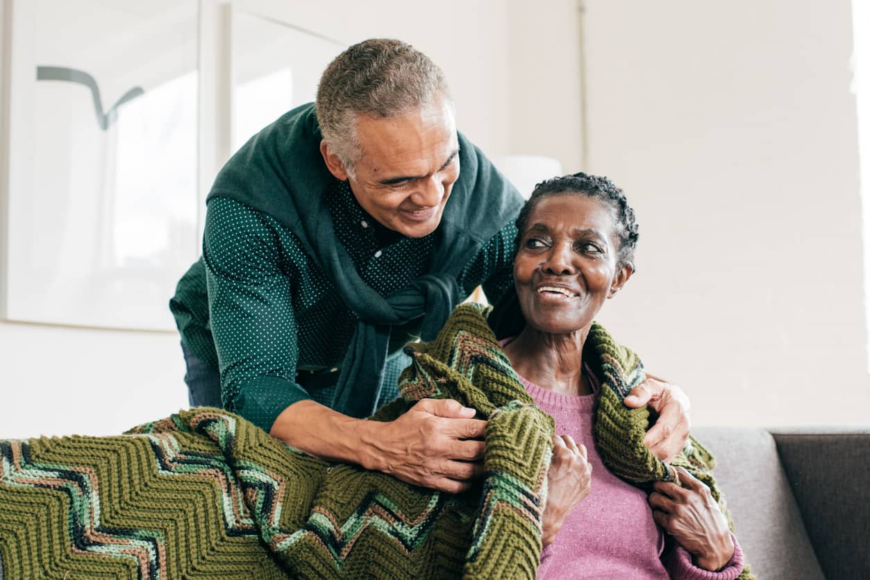 Son wrapping his mother in a knitted blanket as they smile at each other lovingly.