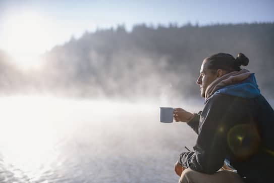 A male hider drinking coffee at sunrise at the edge of a steaming lake.