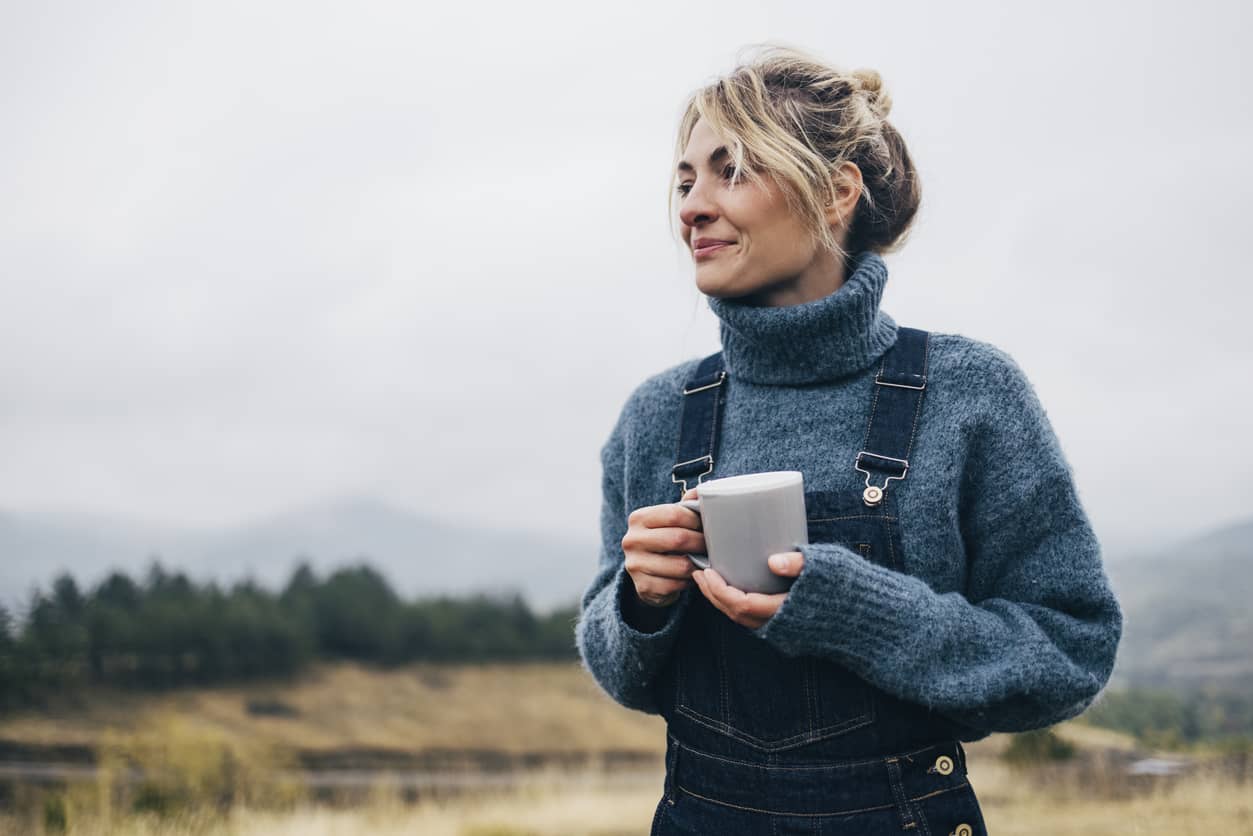 A young woman in overhauls standing in a field mindfully. with a cup of coffee.