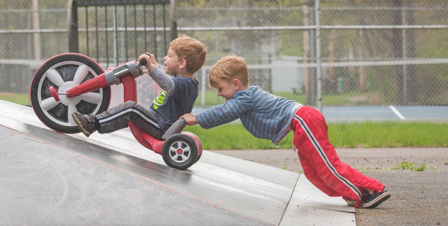 A high energy child pushing his brother up a hill at a skatepark.