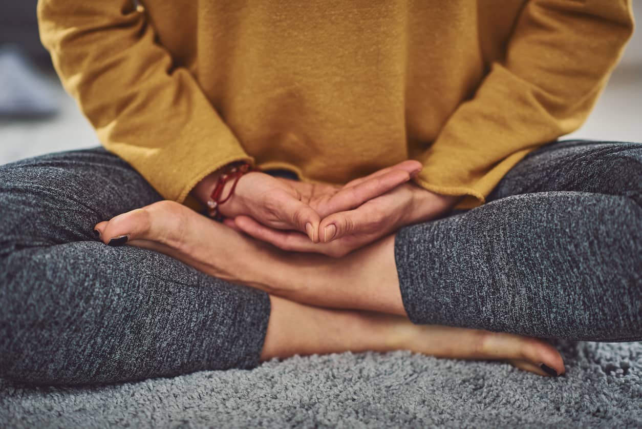 A woman in a bright yellow shirt and black yoga pants is sitting cross-legged taking deep breaths to calm her anxiety.