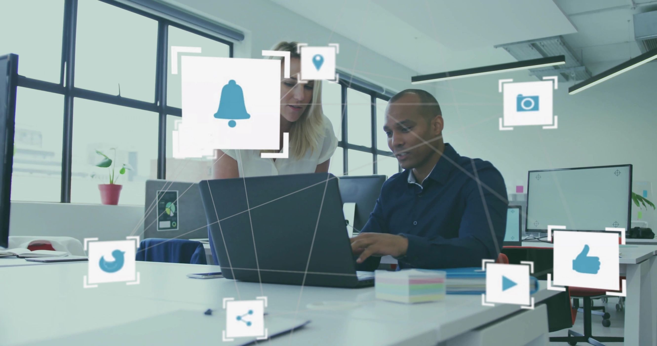 Collaborating coworkers in business attire leaning over laptop at office with digital icons bombarding them.