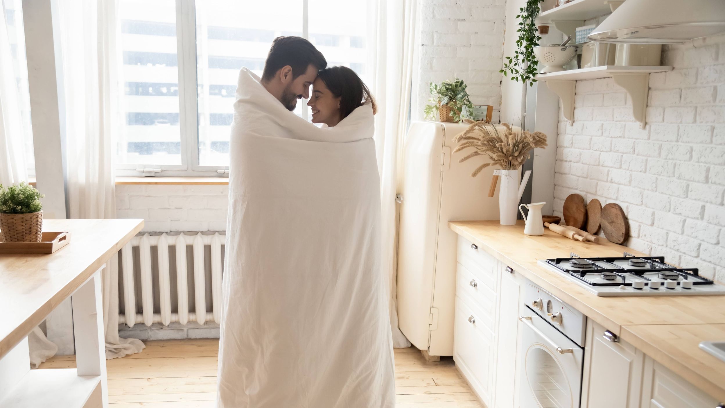 Couple standing in their kitchen face-to-face wrapped in blanket.