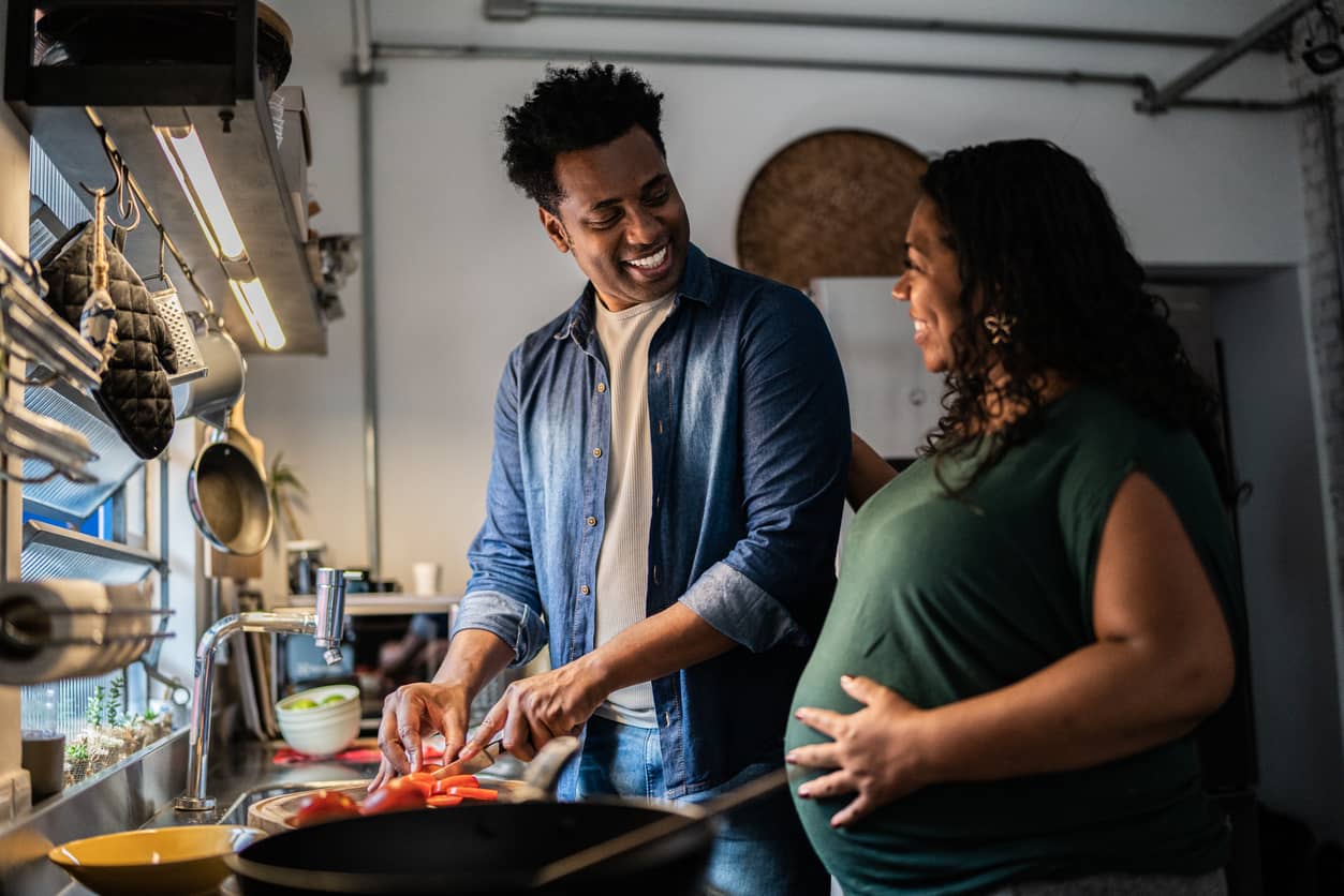 A black pregnant woman is cooking dinner in the kitchen with her husband.