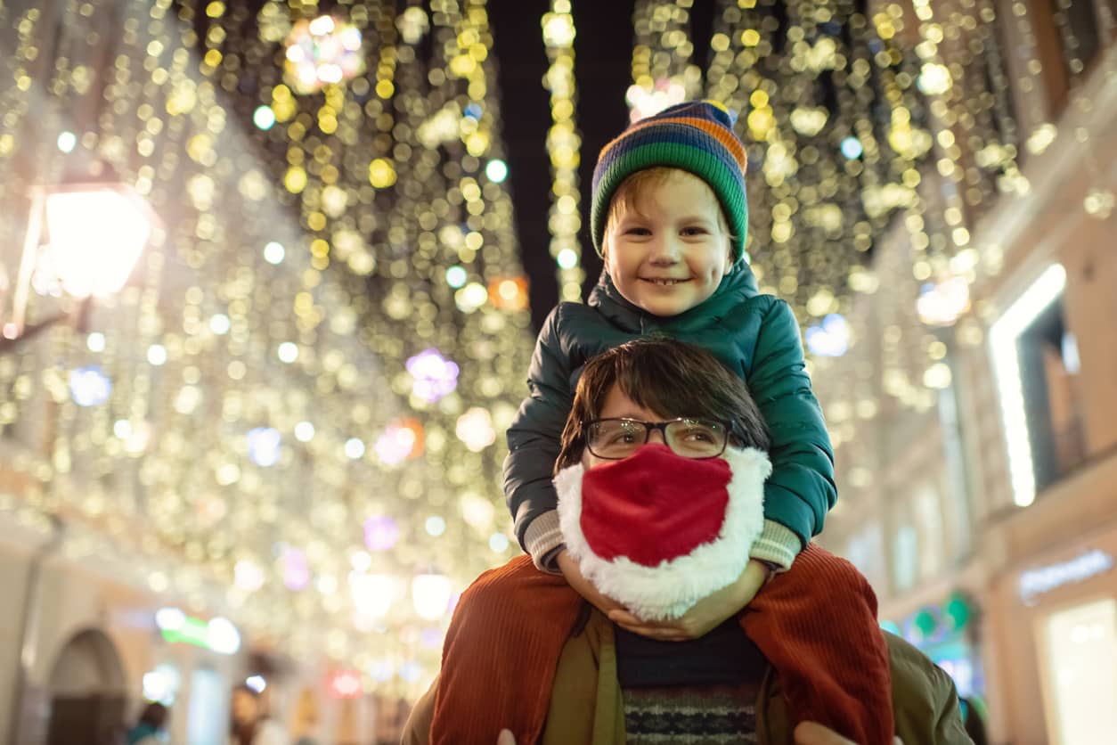 Family enjoying Christmas lights; small boy on the shoulders of his mother who is wearing a red Santa beard.