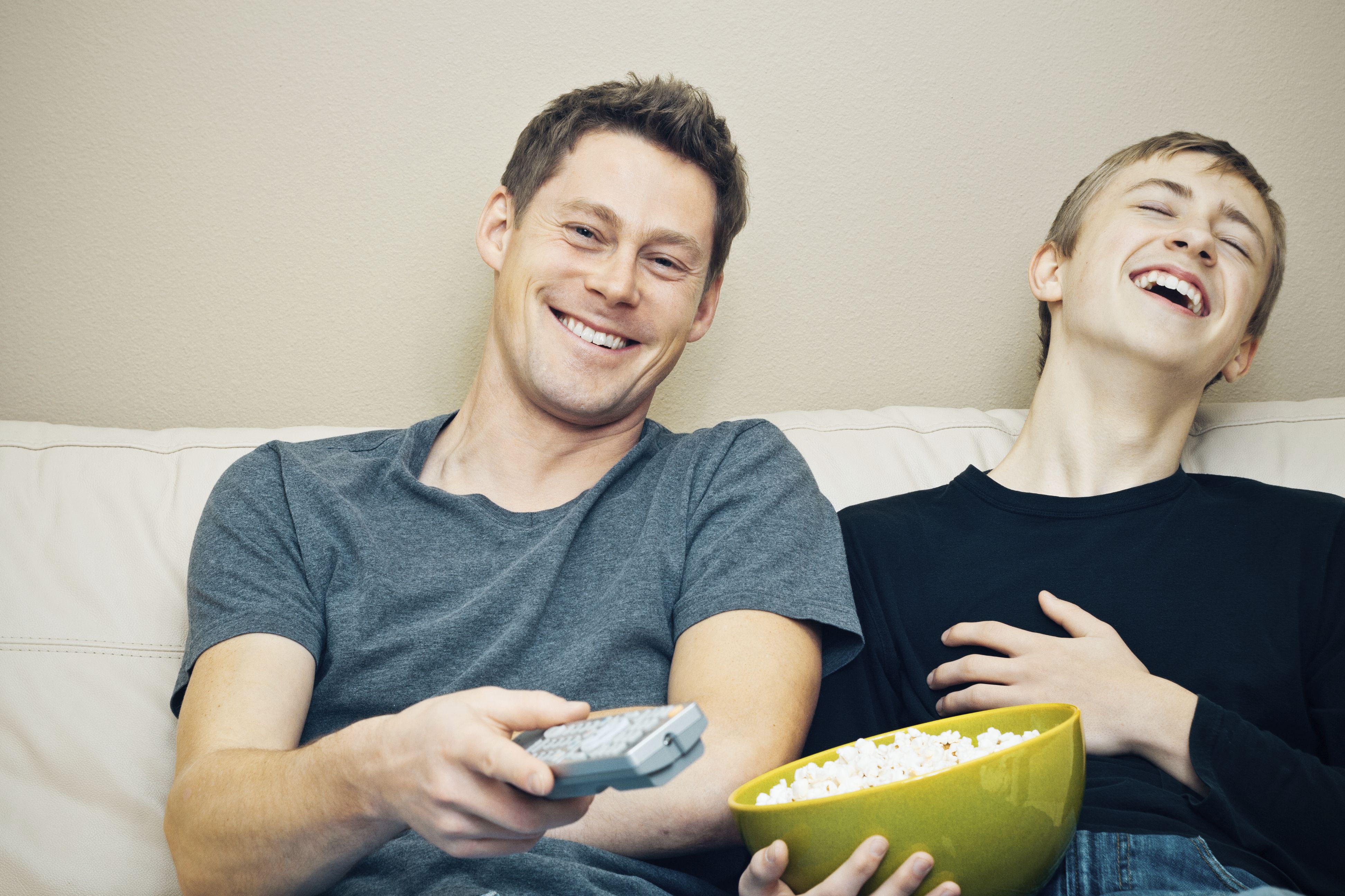 Father and teenage son laughing as they watch TV together.