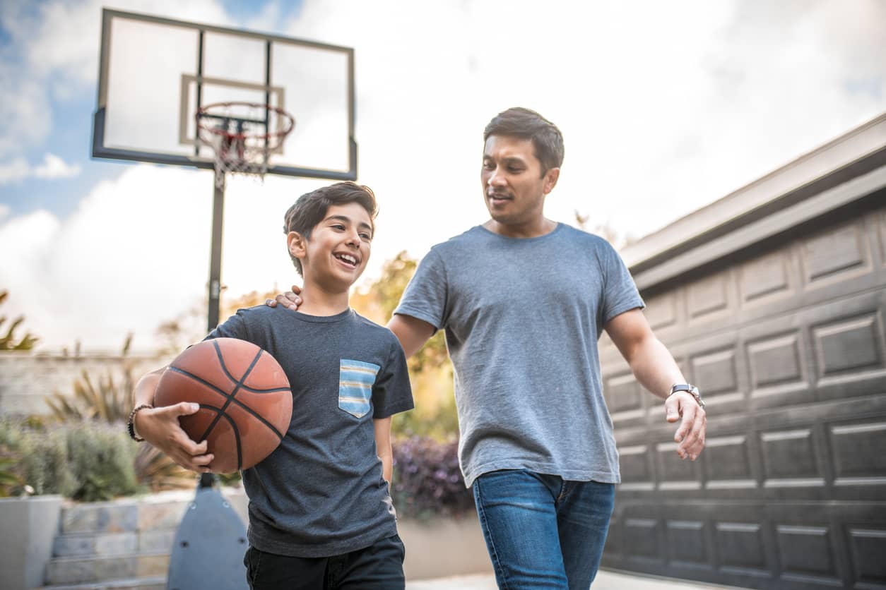 A young teenage boy in a blue T-shirt is holding a basketball as he and his father talk and walk away from the hoop after playing basketball.