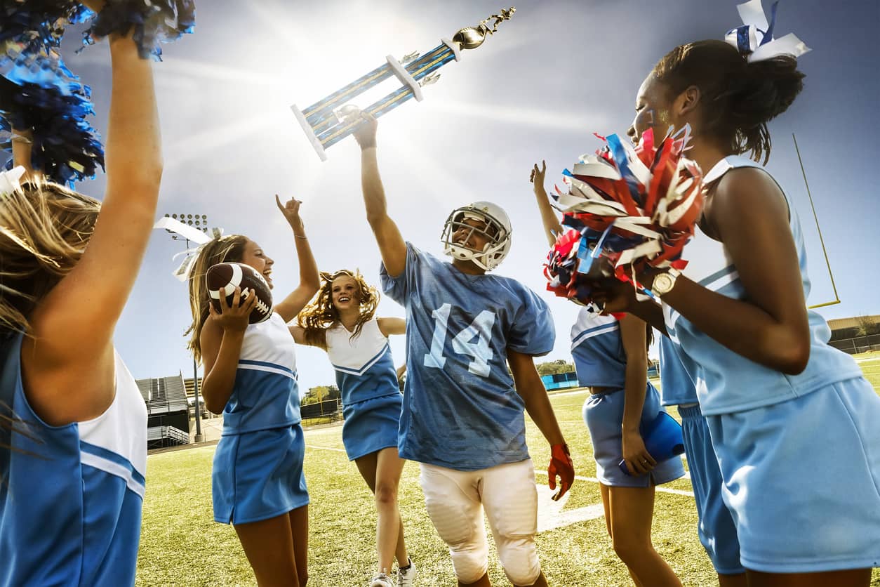 Football player holding up a trophy with a triumphant shout surrounded by cheerleaders.