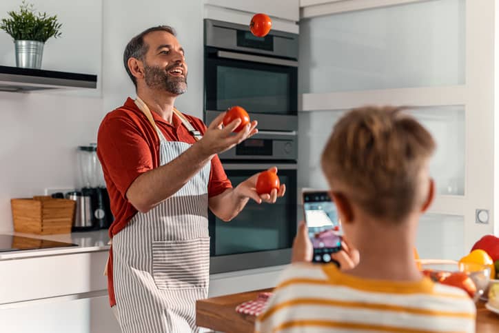 A father juggling tomatoes in the kitchen while his son videos him with a smart phone.