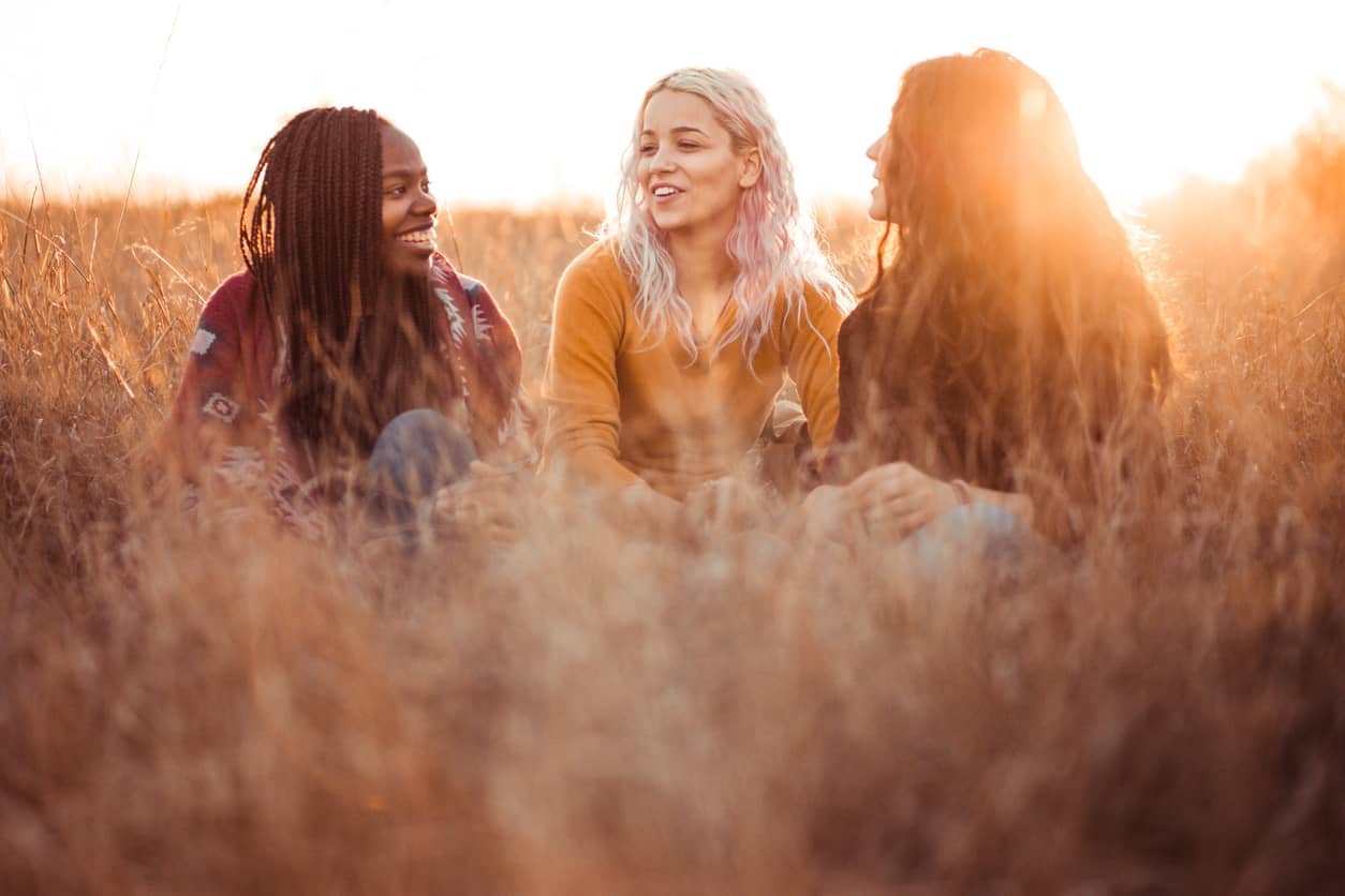 Three women sitting and talking in a field with a golden sun in the background.