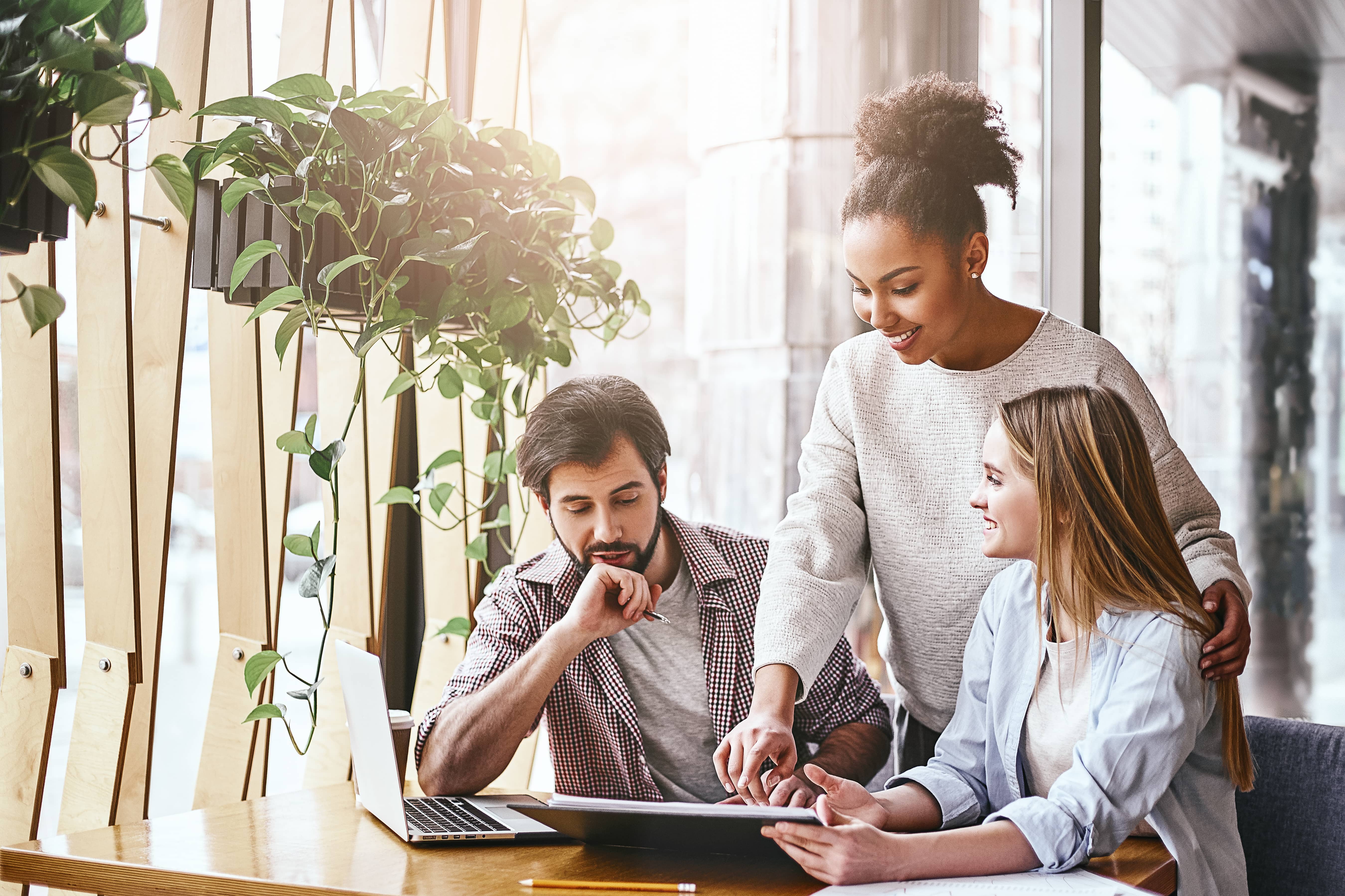 Emotional intelligence at work: a smiling black woman stands behind two employees seated at a computer with a spreadsheet.