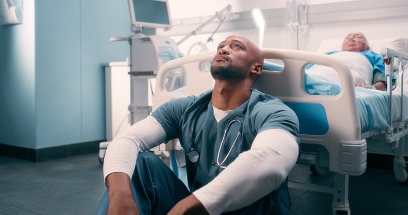 Healthcare provider stressed and exhausted sitting on floor at the foot of a hospital bed.
