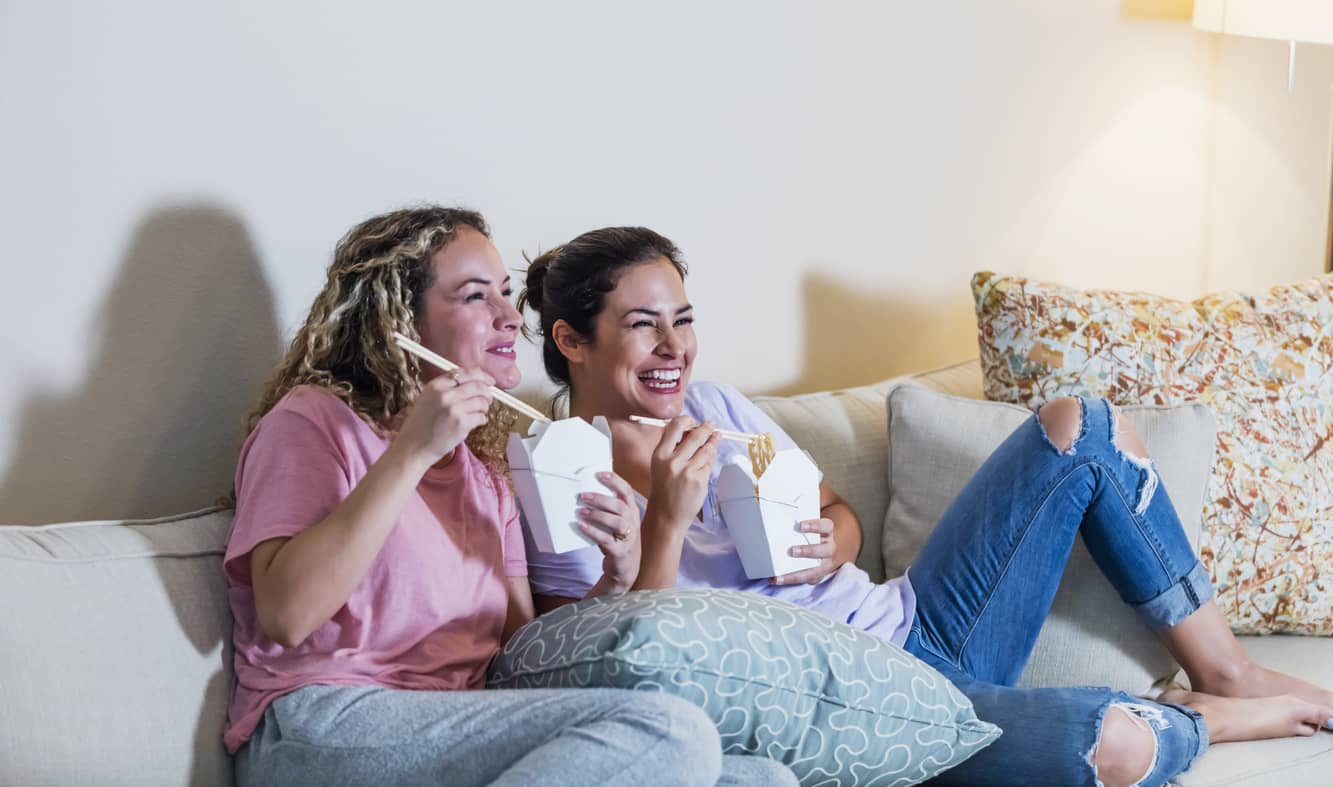 Two hispanic women sitting on sofa eating take-out with chopsticks and watching a movie.