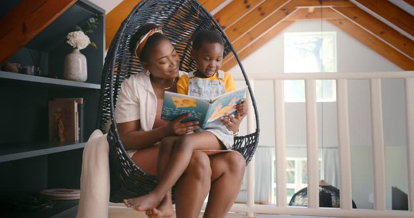 A black mom reading a story to her daughter in a cozy wicker indoor swing.