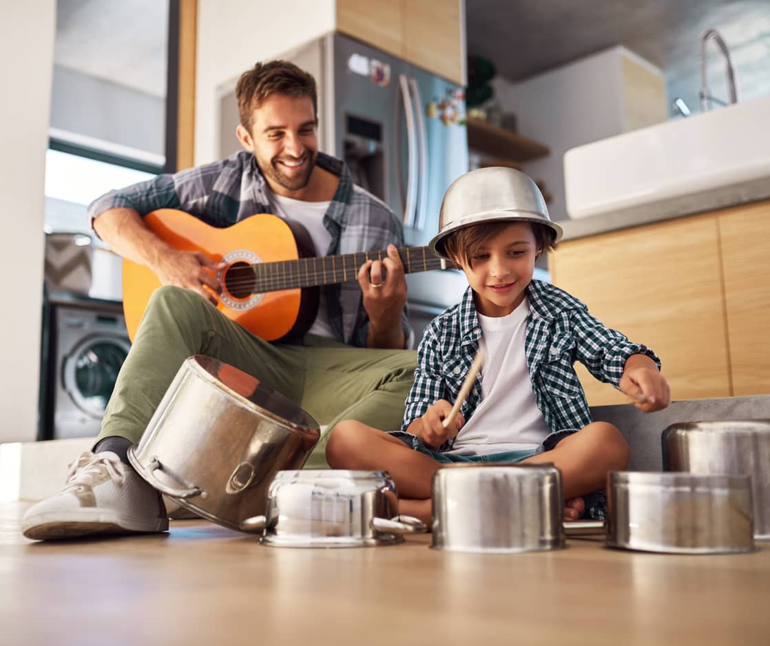 Father playing guitar while his high-energy son bangs the pots and pans joyfully.