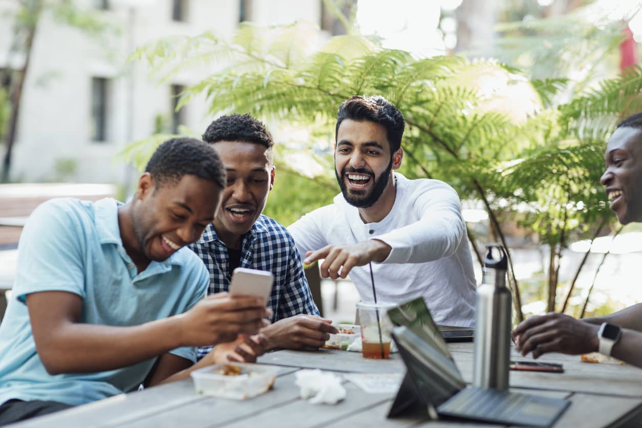 Three men at lunch laughing together while viewing a TikTok video on a smartphone.