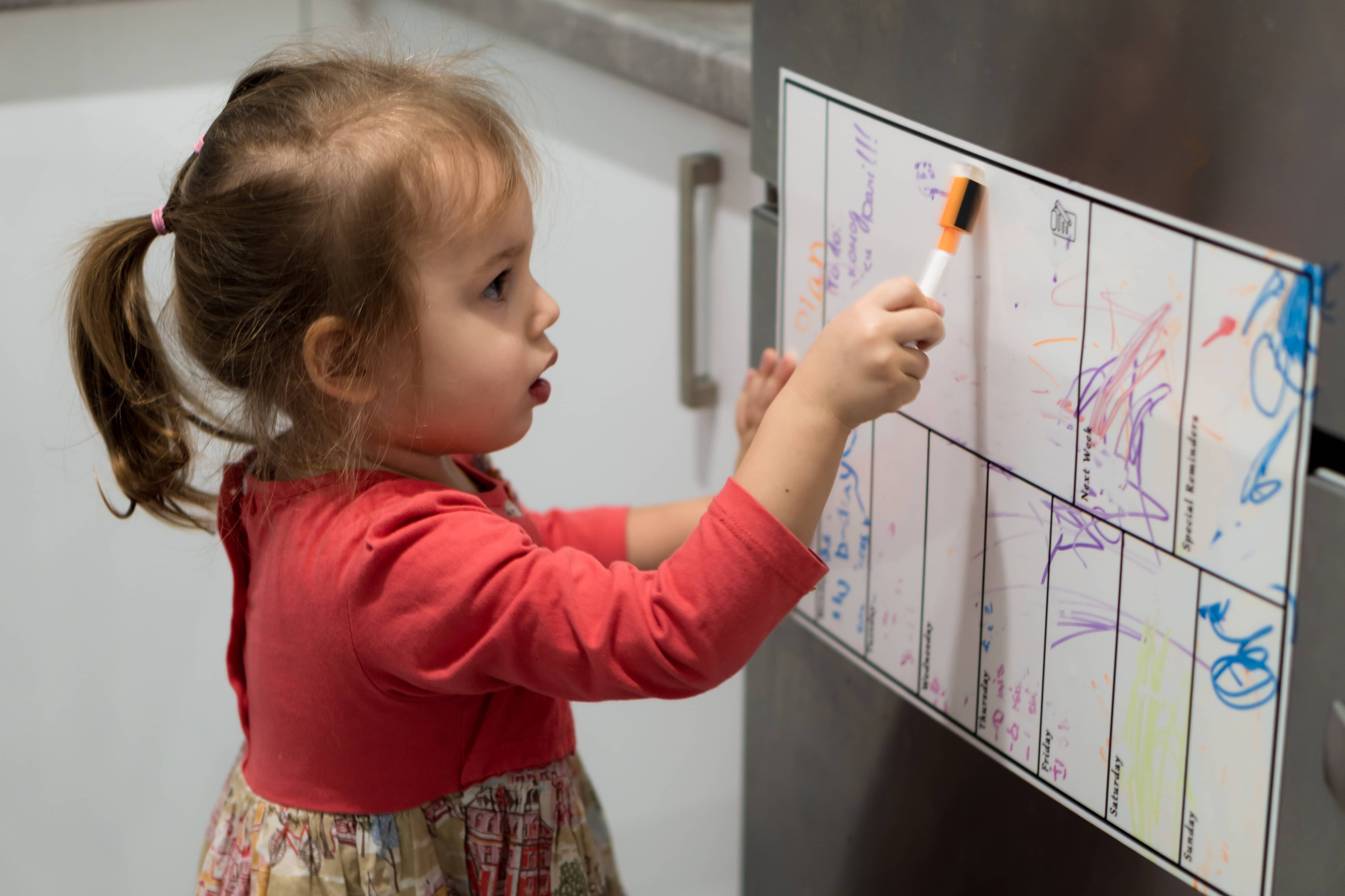 Little girl drawing with markers on a refrigerator calendar.