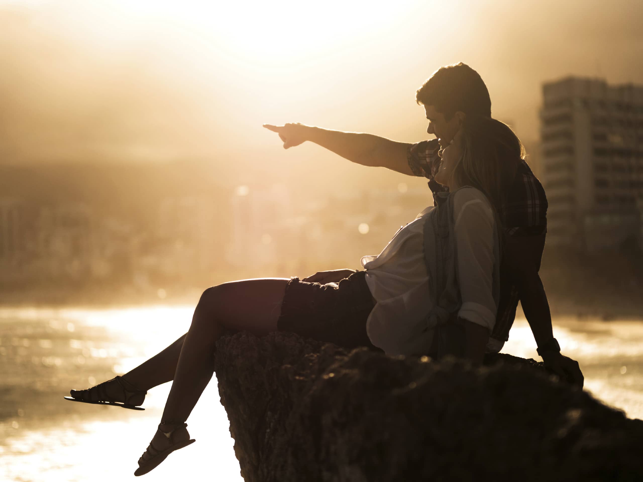 A man pointing out a sailboat to his wife while they sit on top of a large rock.