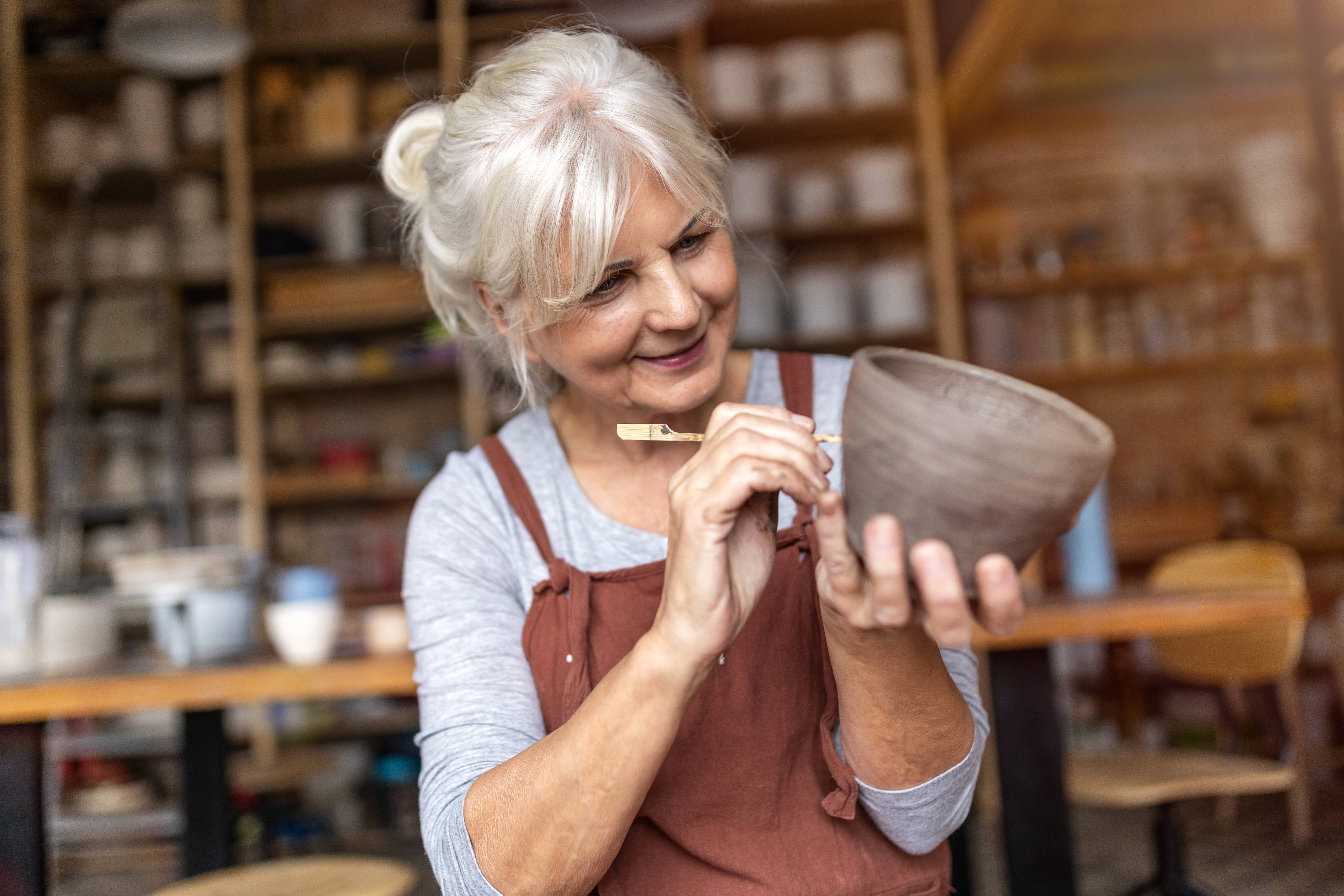 An older woman with a rust apron painting a homemade pot; pottery is one of her passions.