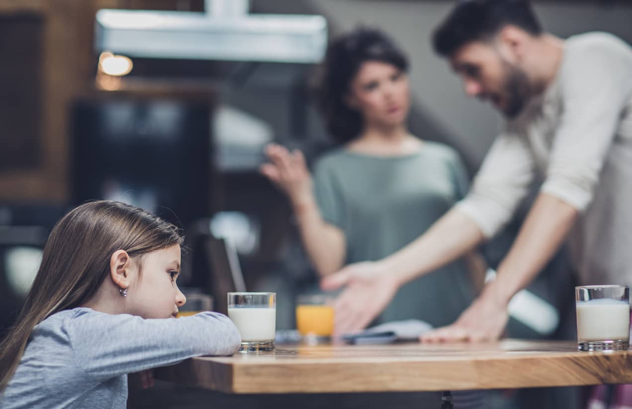 Parenting arguing over differences in parenting styles with sad girl left alone at breakfast table.