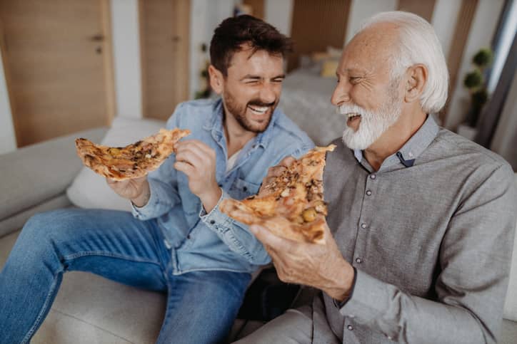 A father and adult son having pizza together and laughing.