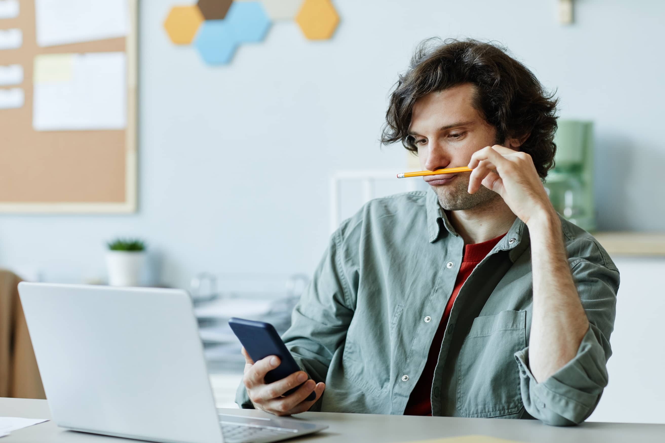 Young man playing with pencil and using his smartphone at work to distract from procrastination.