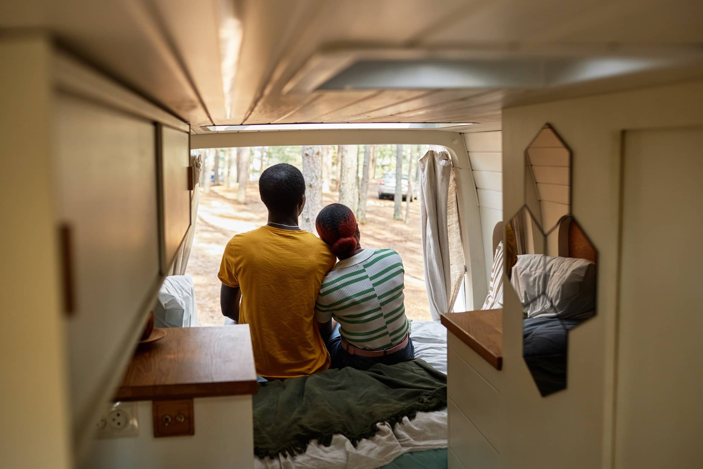 Rear view of black young couple enjoying nature together and embracing while sitting on bed in their travel van.