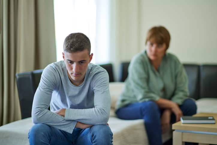 A sad teenage boy in trouble with a mother looking at him in the background.