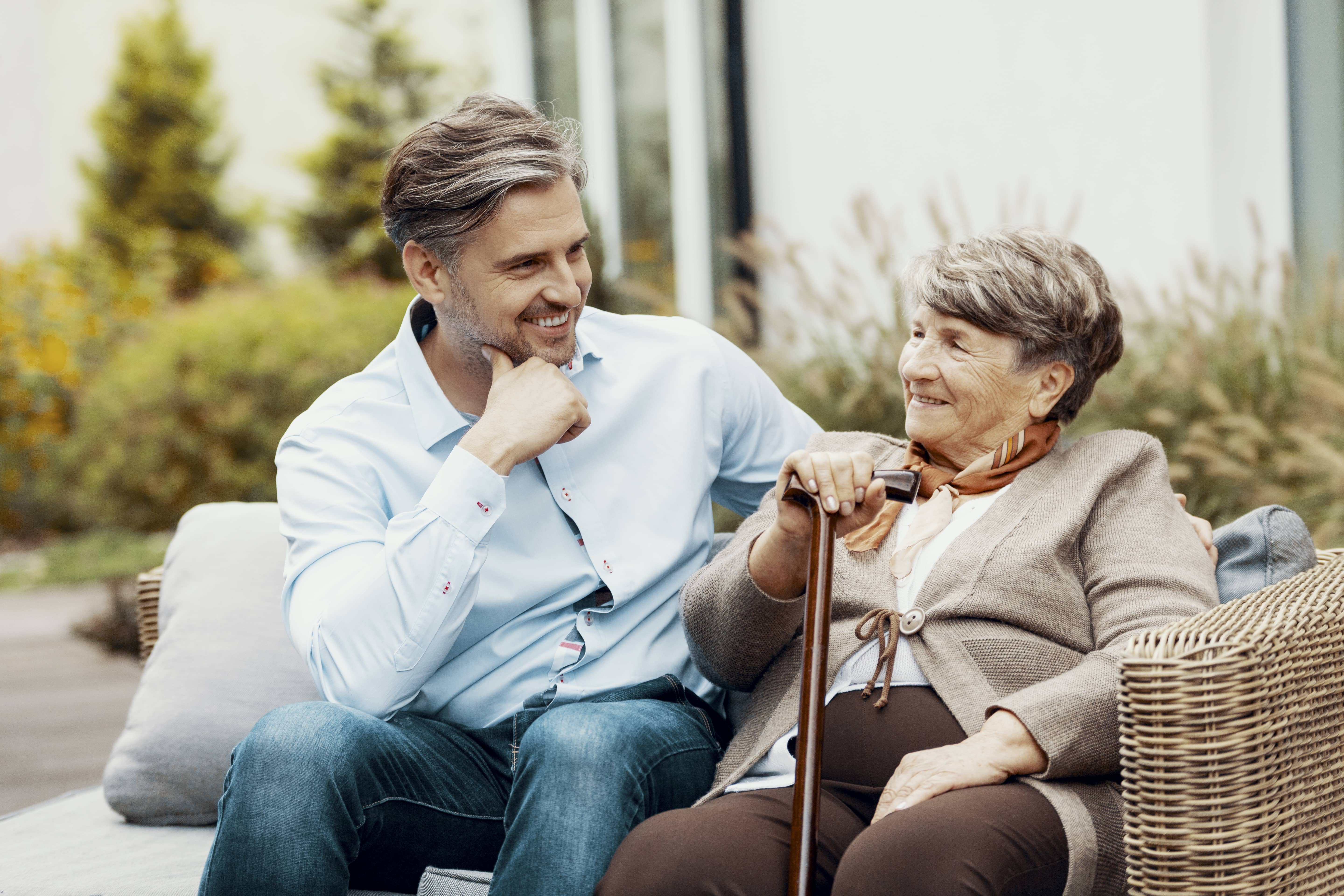 Senior woman with cane looking at grown son as they sit on a wicker bench together.