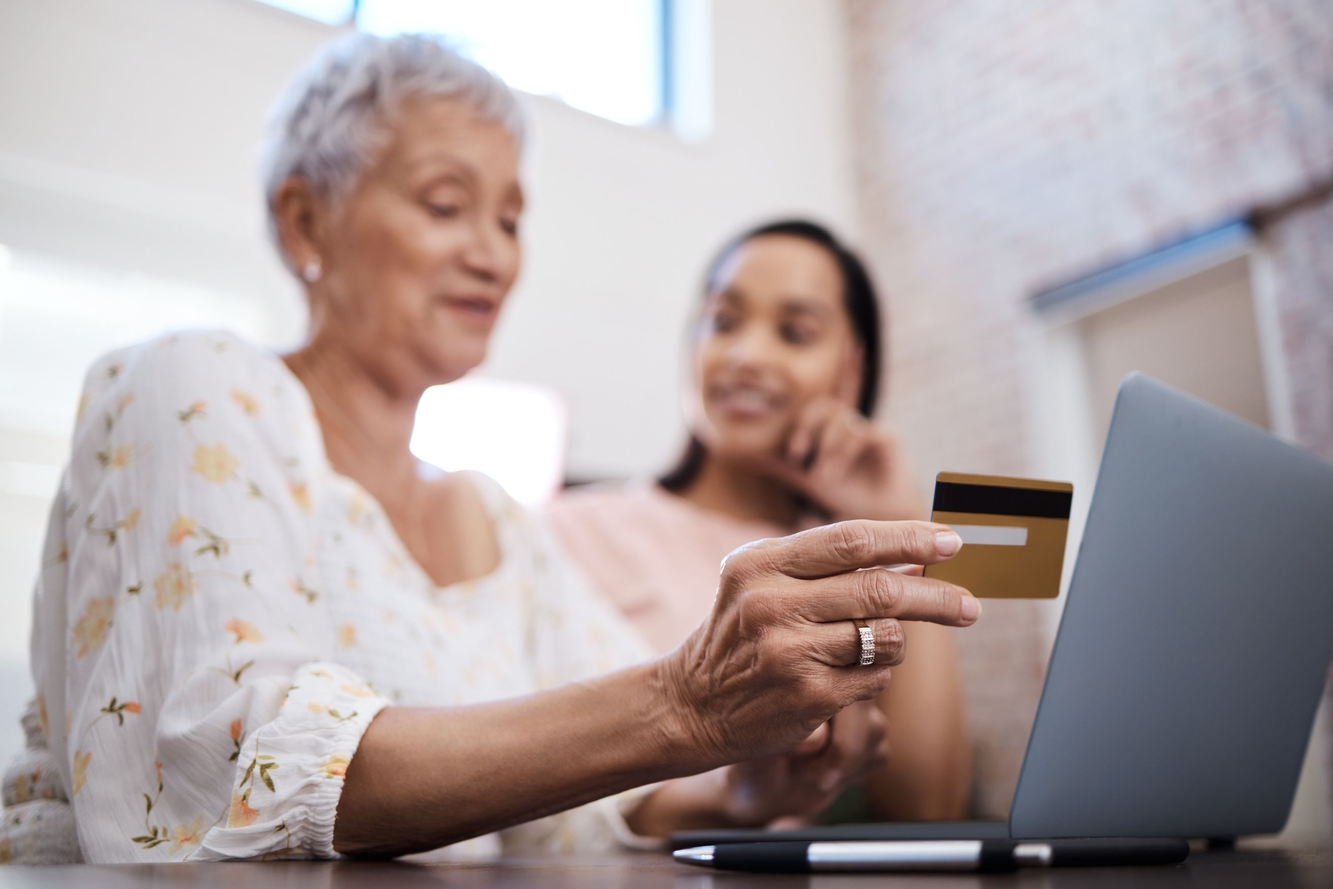 A mother using a laptop and credit card with her adult daughter smiling in the background.