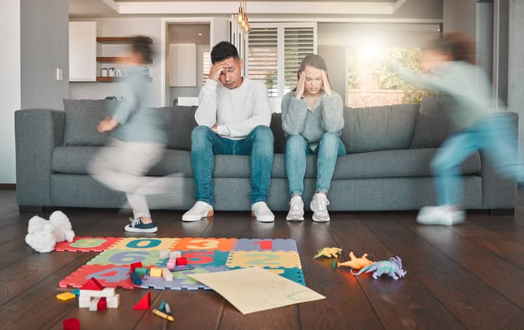 Two young parents looking frustrated and tired while their children run around at home.