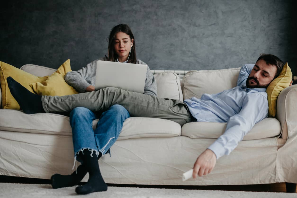 An exhausted man sleeping on his wife's lap as she works on her computer.