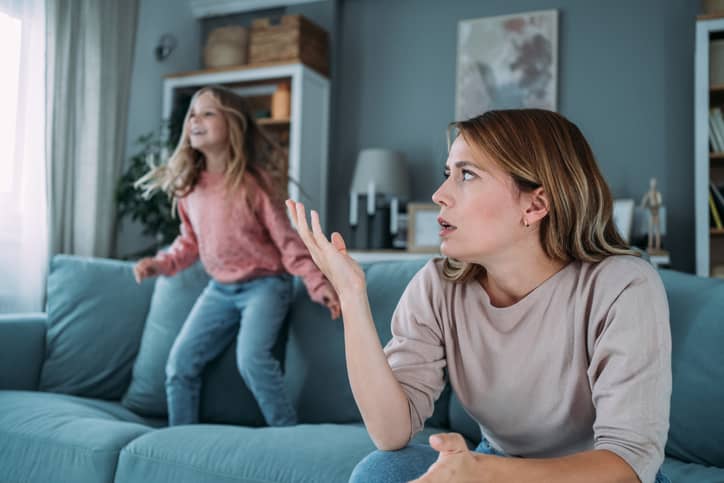 Stressed mother with her preschool daughter jumping on the couch.