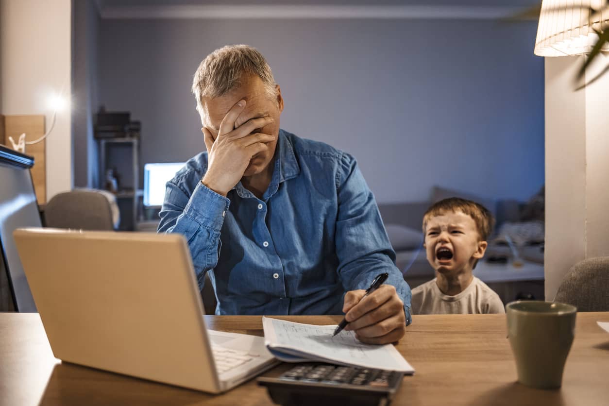 Stressed man with hand over his forehead with his two-year-old son crying next to him..