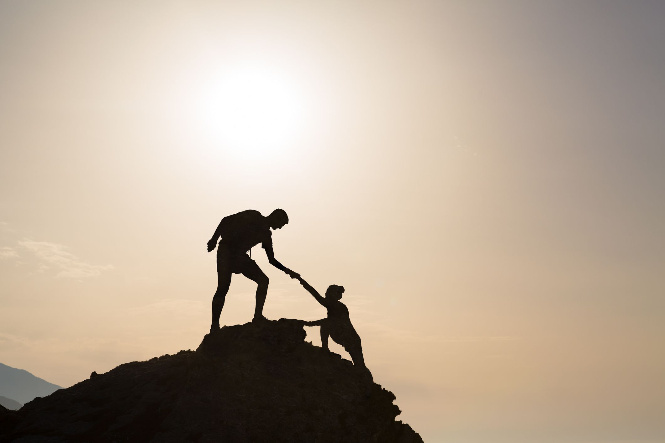 A silhouette of a person with outreached arm and hand helping another climb a mountain.