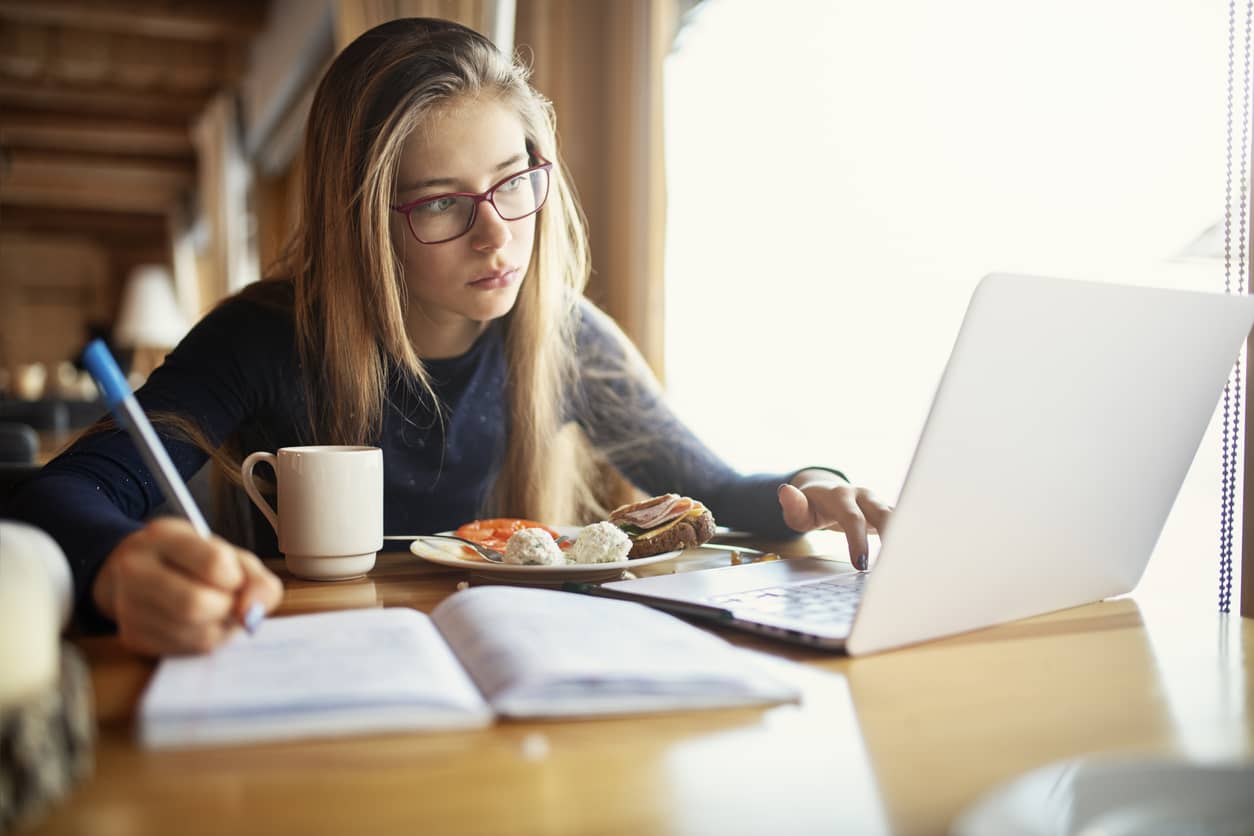 A teenage girl with long blonde hair focusing on her homework, writing in a notebook with her right hand and typing on a computer with her left hand.