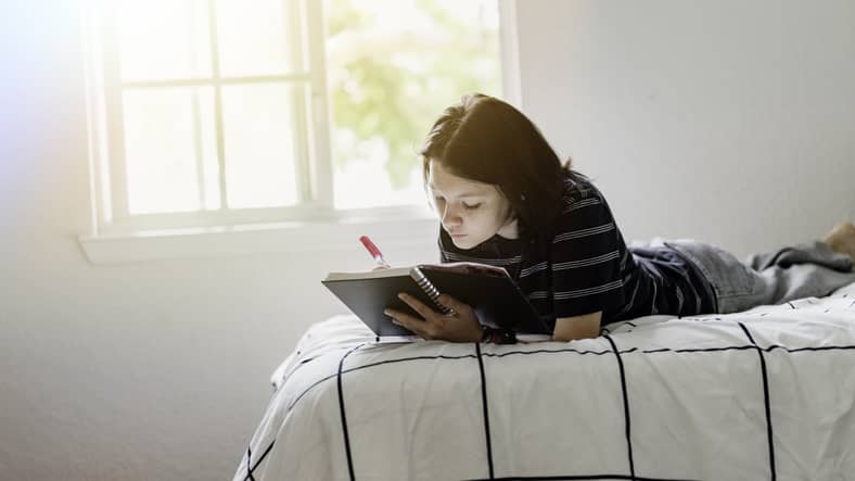 A transgender nonbinary teenager journaling on a bed while sorting through the changes.