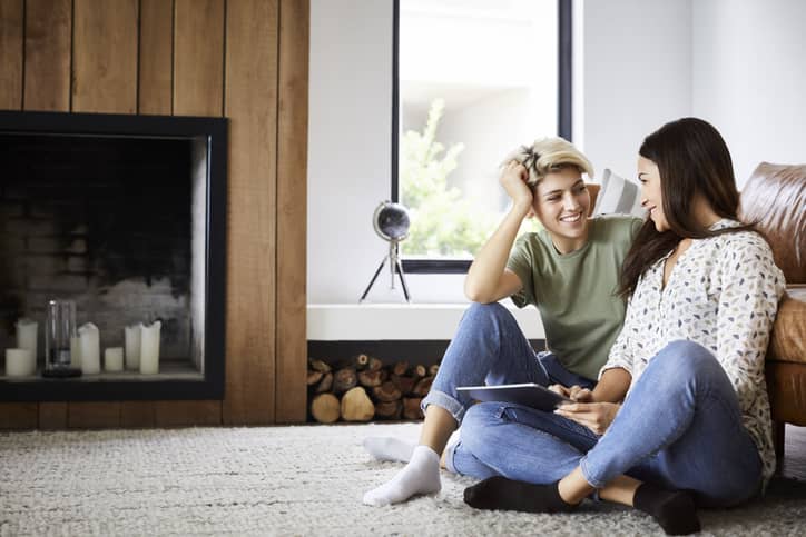 Two women talking in the living room leaning up against the couch.