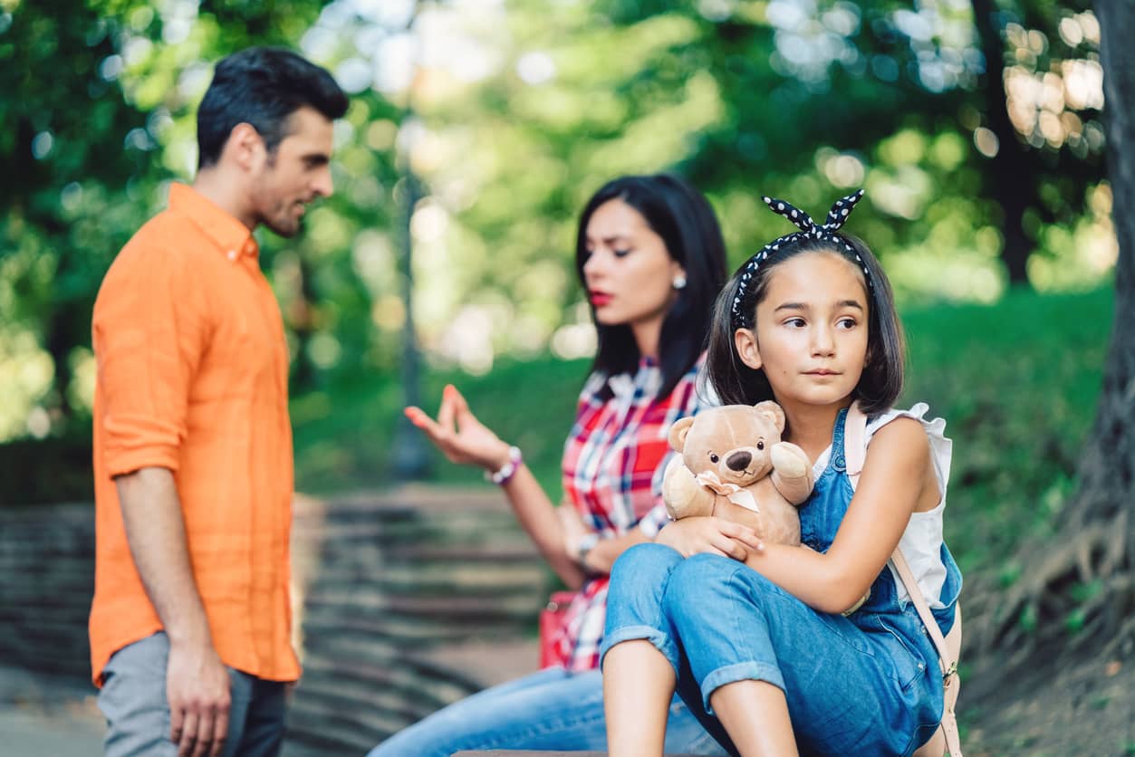 Couple arguing in a park after a divorce while their elementary daughter is listening with a sad face.