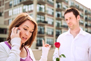 A woman on her phone unaware that her partner is holding out a rose to give her
