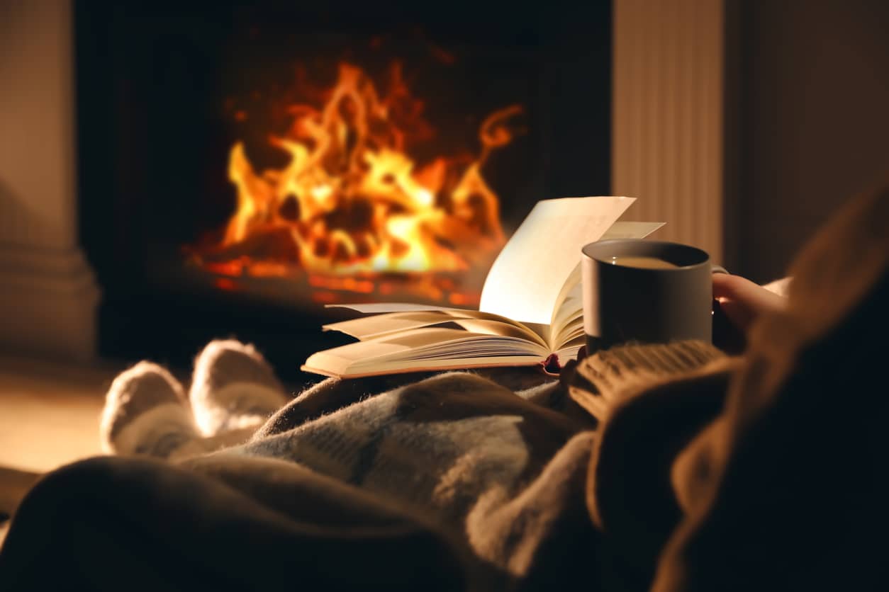 A woman sitting by a fire journaling with a cup of coffee in her hand.