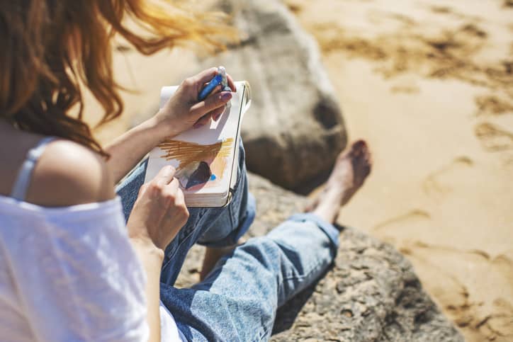 Young adult woman art journaling on a trip to the beach.